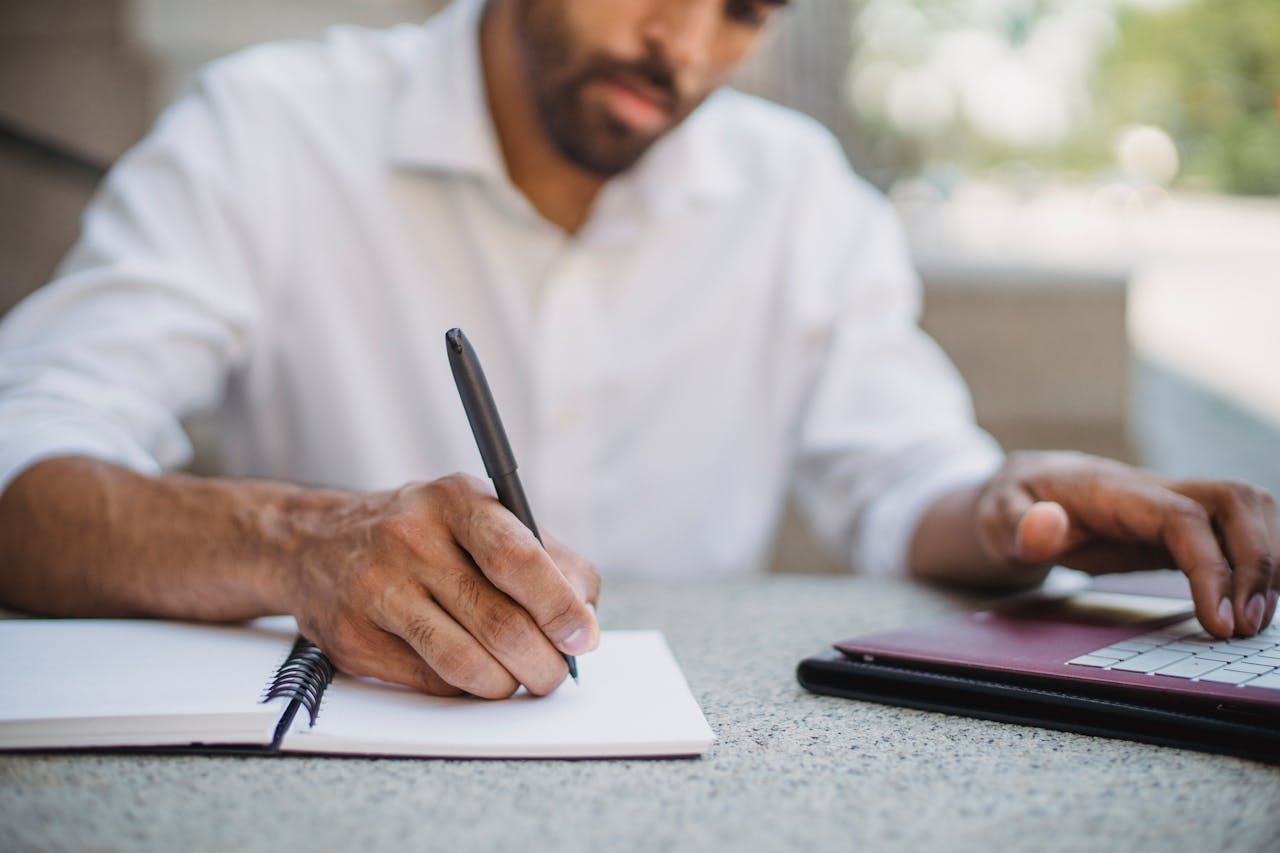 Shallow Focus Photo of a Person Writing on a Notebook while Using His Laptop